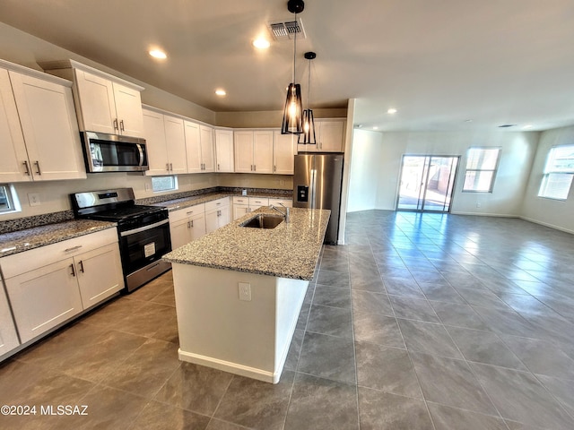 kitchen with a kitchen island with sink, stainless steel appliances, dark stone counters, decorative light fixtures, and white cabinetry