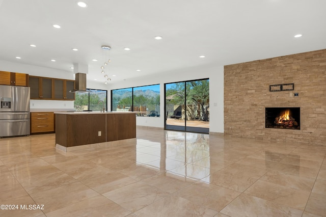 kitchen with hanging light fixtures, a fireplace, an island with sink, island range hood, and stainless steel fridge with ice dispenser