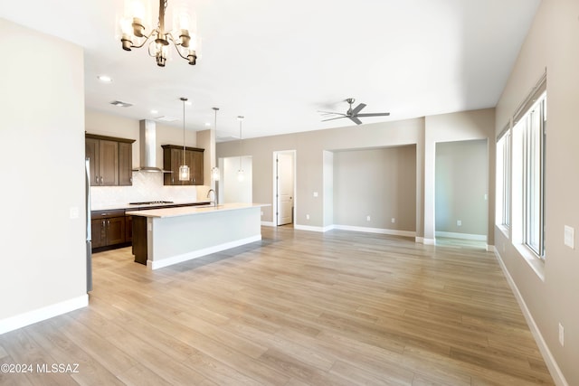 kitchen featuring a kitchen island with sink, wall chimney range hood, hanging light fixtures, light wood-type flooring, and ceiling fan with notable chandelier