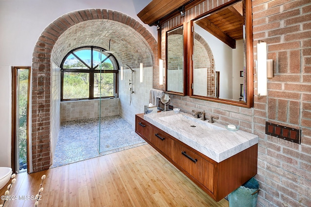 bathroom featuring a tile shower, toilet, vanity, hardwood / wood-style flooring, and brick wall