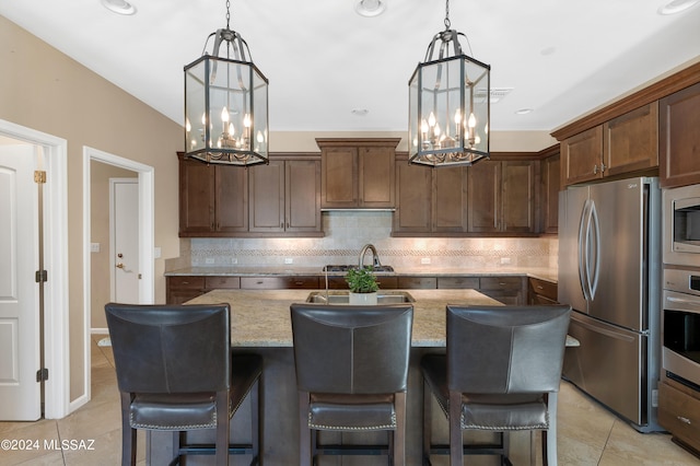 kitchen with tasteful backsplash, appliances with stainless steel finishes, an island with sink, pendant lighting, and light stone counters