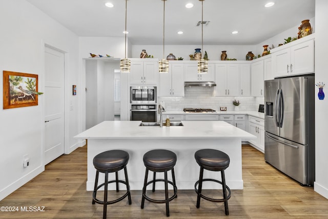 kitchen with white cabinetry, stainless steel appliances, an island with sink, pendant lighting, and light wood-type flooring