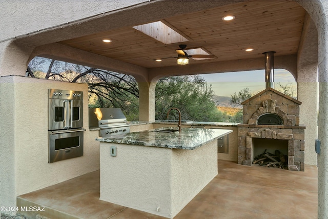 patio terrace at dusk with ceiling fan, area for grilling, sink, and an outdoor stone fireplace