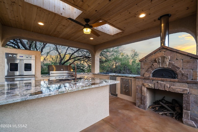 patio terrace at dusk with area for grilling, an outdoor stone fireplace, grilling area, and ceiling fan