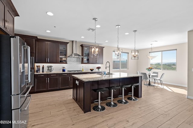 kitchen with wall chimney exhaust hood, an island with sink, sink, appliances with stainless steel finishes, and light hardwood / wood-style floors