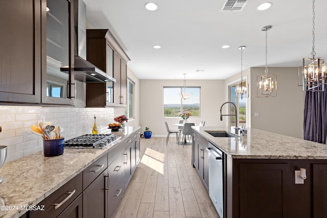 kitchen with wall chimney range hood, an island with sink, sink, appliances with stainless steel finishes, and light hardwood / wood-style floors