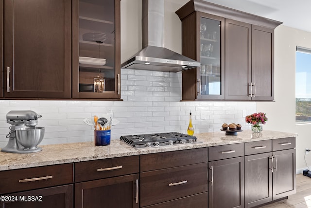 kitchen featuring wall chimney range hood, dark brown cabinetry, stainless steel gas stovetop, light stone counters, and tasteful backsplash