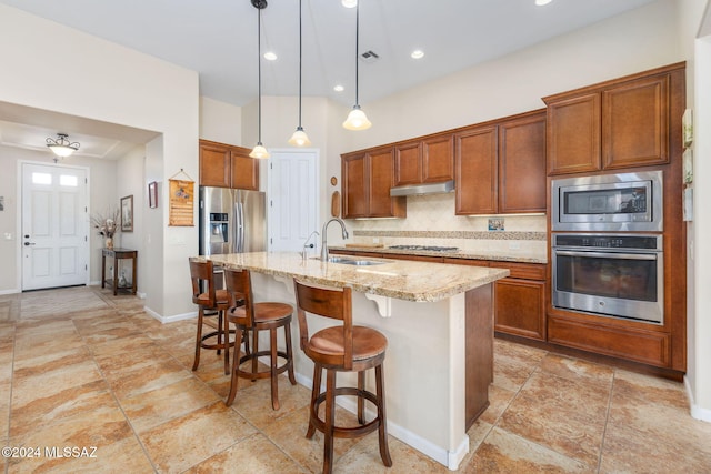 kitchen with light stone counters, stainless steel appliances, sink, pendant lighting, and an island with sink