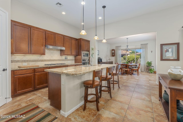 kitchen featuring a kitchen bar, backsplash, stainless steel appliances, decorative light fixtures, and a center island with sink