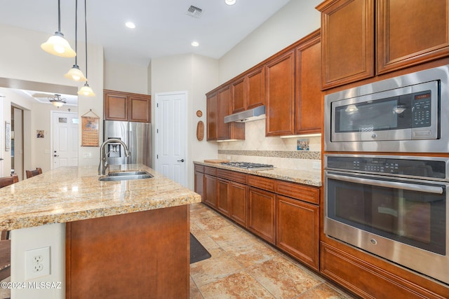 kitchen featuring appliances with stainless steel finishes, light stone counters, sink, a center island with sink, and decorative light fixtures