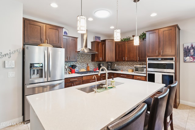 kitchen with sink, hanging light fixtures, wall chimney range hood, a center island with sink, and appliances with stainless steel finishes