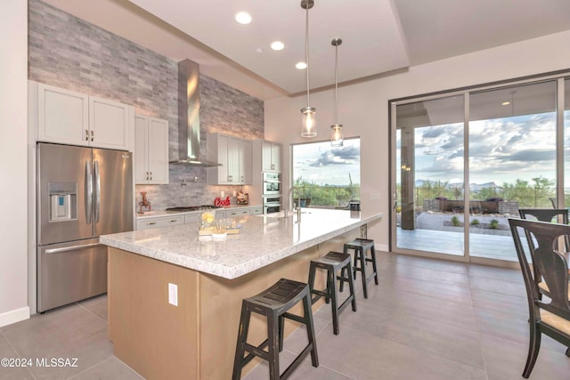 kitchen featuring stainless steel appliances, a center island with sink, light stone counters, white cabinets, and wall chimney exhaust hood