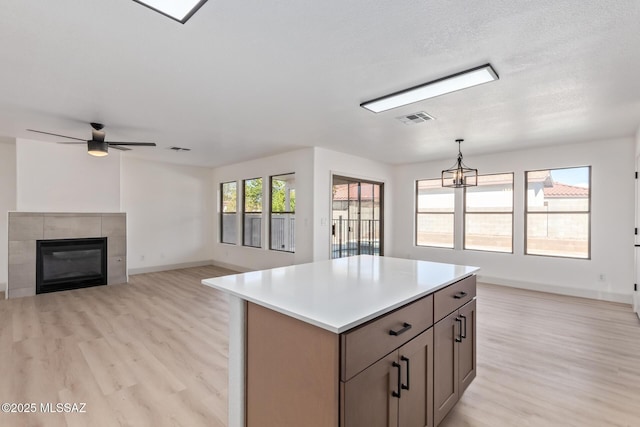 kitchen with pendant lighting, light hardwood / wood-style floors, a kitchen island, a tiled fireplace, and ceiling fan with notable chandelier