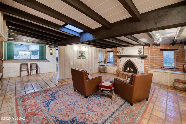 tiled living room featuring wooden walls, a healthy amount of sunlight, beam ceiling, and a brick fireplace