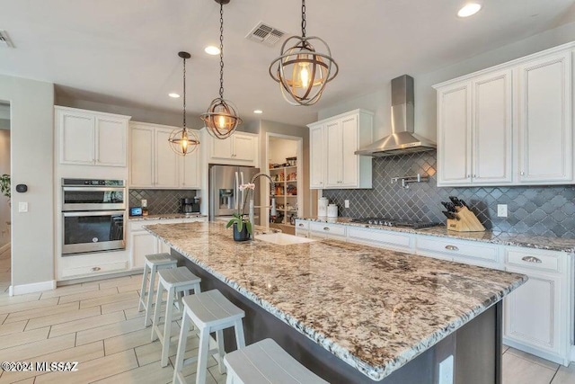 kitchen with white cabinetry, stainless steel appliances, wall chimney exhaust hood, pendant lighting, and a kitchen island with sink