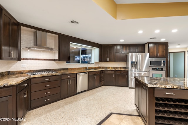 kitchen with sink, stainless steel appliances, wall chimney range hood, dark stone counters, and dark brown cabinets