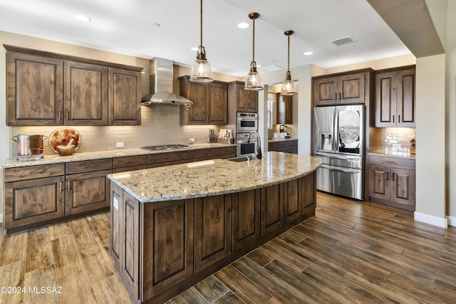 kitchen with hanging light fixtures, stainless steel appliances, wall chimney range hood, tasteful backsplash, and a center island with sink