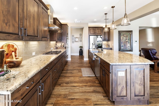 kitchen featuring pendant lighting, a center island with sink, wall chimney range hood, decorative backsplash, and appliances with stainless steel finishes