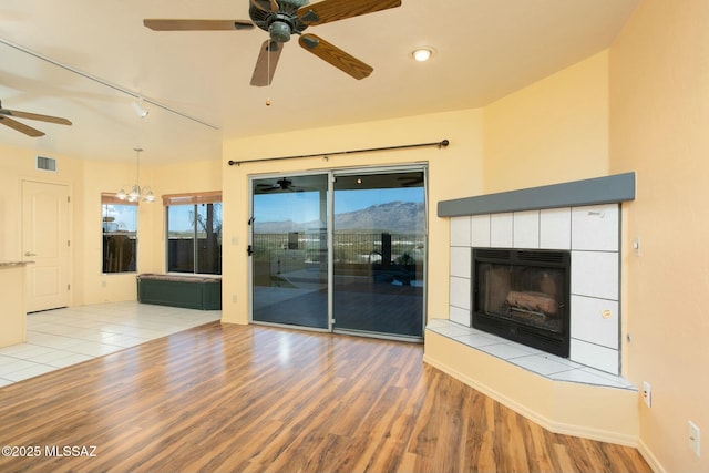 unfurnished living room with ceiling fan with notable chandelier, light hardwood / wood-style floors, rail lighting, and a tiled fireplace