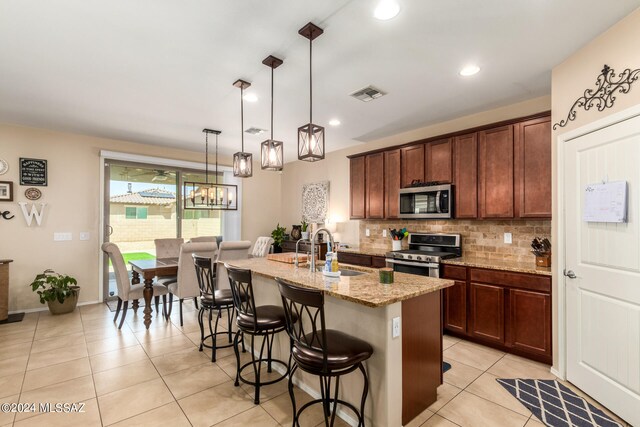 kitchen with an island with sink, appliances with stainless steel finishes, light stone countertops, pendant lighting, and sink