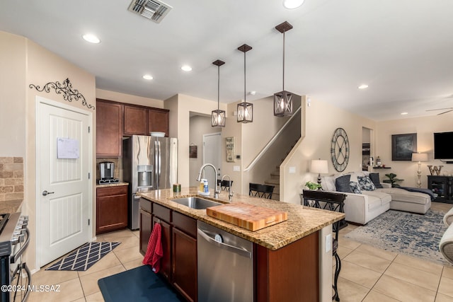 kitchen with stainless steel appliances, a center island with sink, sink, light stone countertops, and pendant lighting