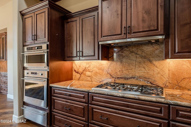 kitchen featuring light stone counters, under cabinet range hood, stainless steel appliances, dark brown cabinets, and a warming drawer