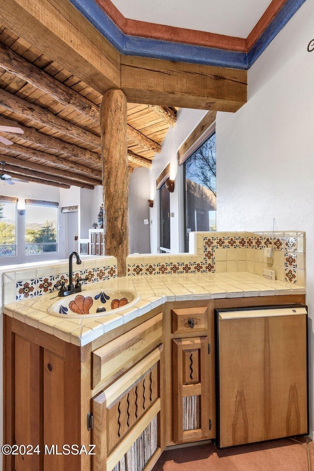 kitchen featuring beam ceiling, tile countertops, brown cabinetry, wood ceiling, and a sink