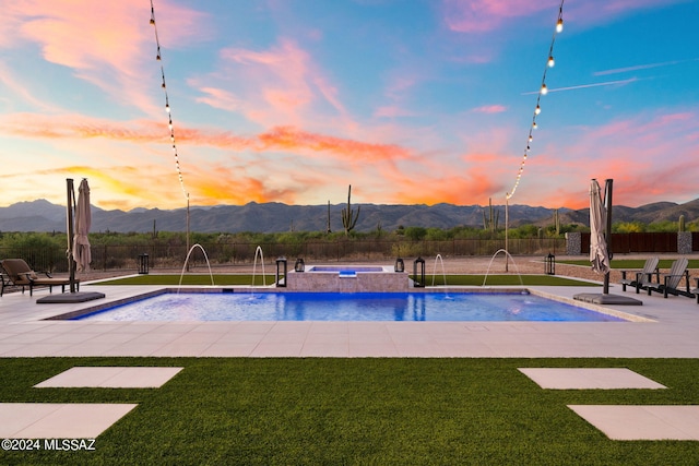 pool at dusk with a lawn, a mountain view, fence, and an in ground hot tub