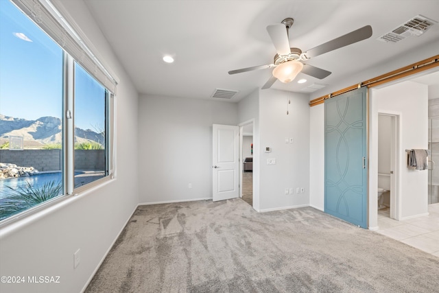 unfurnished bedroom with light carpet, a barn door, ceiling fan, and connected bathroom