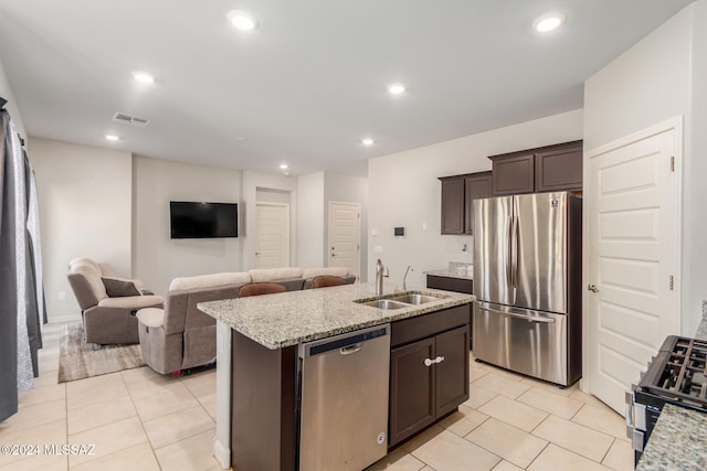 kitchen with appliances with stainless steel finishes, dark brown cabinets, light stone countertops, sink, and an island with sink