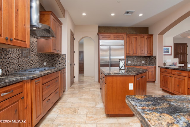 kitchen with wall chimney exhaust hood, backsplash, dark stone counters, a kitchen island, and appliances with stainless steel finishes