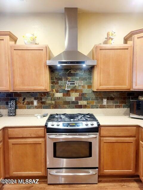 kitchen featuring wall chimney exhaust hood, stainless steel gas range oven, light brown cabinetry, and backsplash