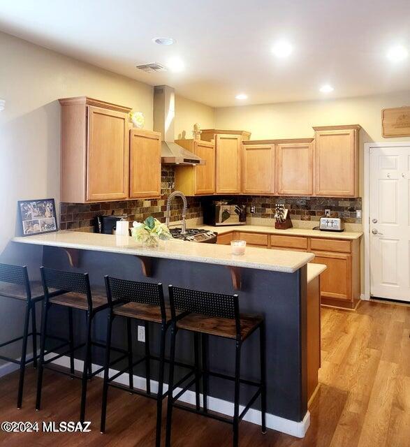 kitchen with wall chimney range hood, kitchen peninsula, tasteful backsplash, a kitchen breakfast bar, and light hardwood / wood-style floors