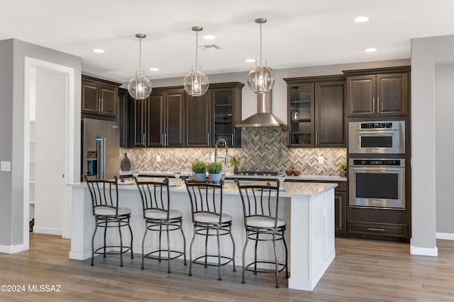 kitchen with wall chimney range hood, a kitchen island with sink, hanging light fixtures, dark brown cabinets, and stainless steel appliances