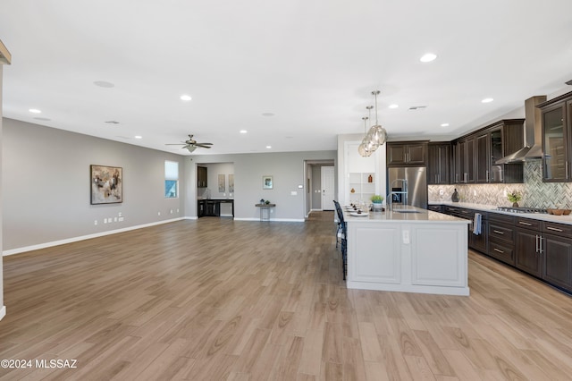 kitchen featuring light hardwood / wood-style floors, a center island with sink, appliances with stainless steel finishes, dark brown cabinets, and wall chimney range hood