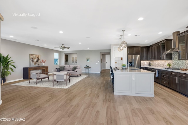 kitchen with appliances with stainless steel finishes, decorative light fixtures, a kitchen island with sink, dark brown cabinetry, and wall chimney range hood