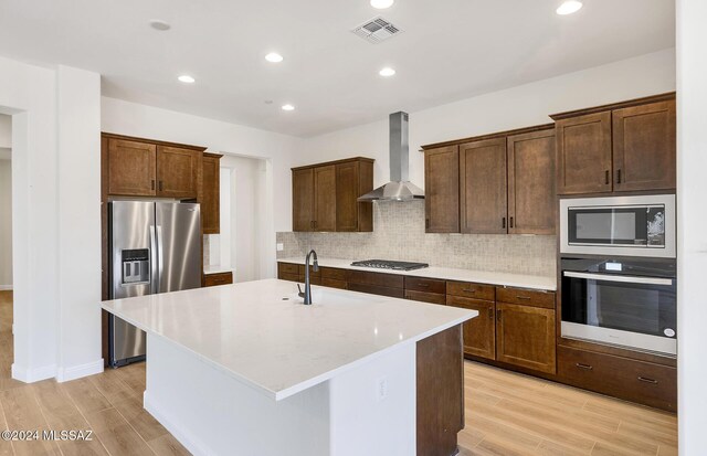 kitchen with a center island with sink, sink, wall chimney exhaust hood, light wood-type flooring, and appliances with stainless steel finishes