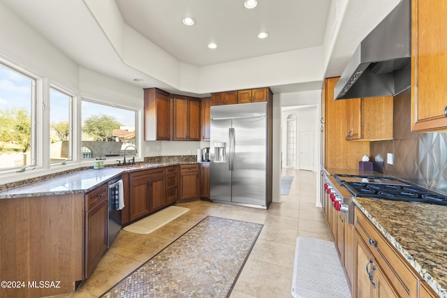 kitchen with dark stone counters, sink, stainless steel appliances, and wall chimney range hood