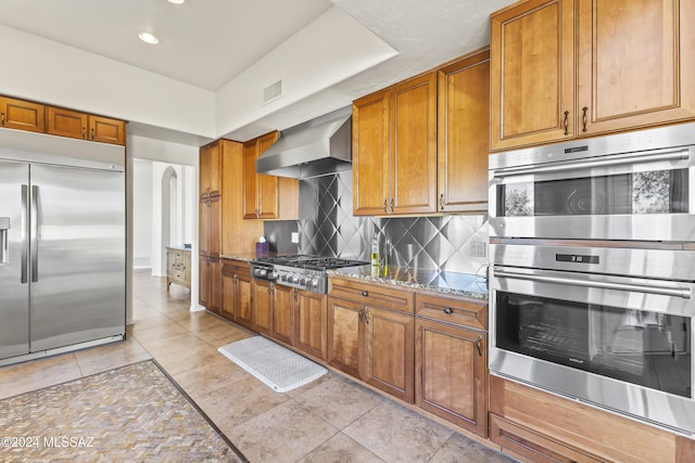 kitchen featuring wall chimney exhaust hood, decorative backsplash, light stone countertops, appliances with stainless steel finishes, and light tile patterned flooring