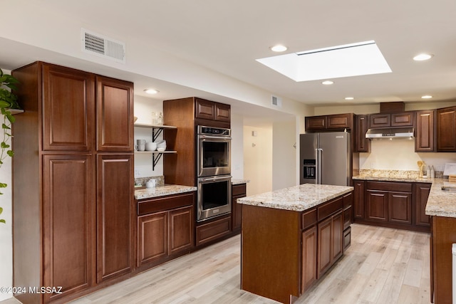 kitchen with appliances with stainless steel finishes, a skylight, a center island, light stone counters, and light wood-type flooring