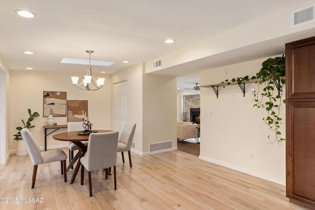 dining space featuring a stone fireplace, ceiling fan with notable chandelier, light wood-type flooring, and a skylight