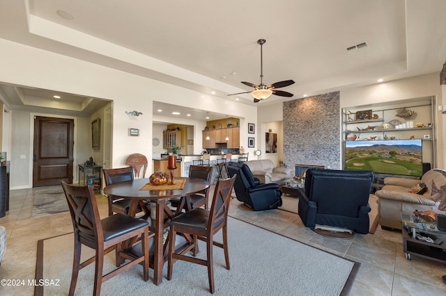 tiled dining room featuring a stone fireplace, ceiling fan, and a tray ceiling