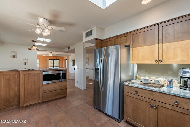 kitchen with a skylight, brown cabinetry, appliances with stainless steel finishes, light stone counters, and a healthy amount of sunlight