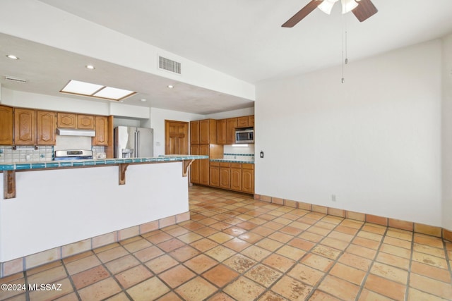 kitchen with appliances with stainless steel finishes, a kitchen bar, decorative backsplash, tile countertops, and ceiling fan