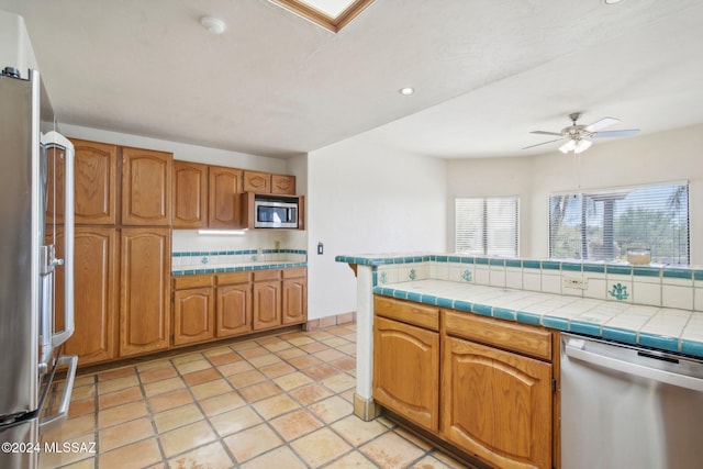 kitchen featuring tile counters, decorative backsplash, light tile patterned floors, ceiling fan, and appliances with stainless steel finishes