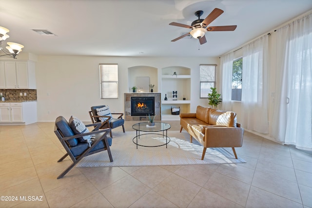 tiled living room featuring a fireplace, built in features, and ceiling fan with notable chandelier