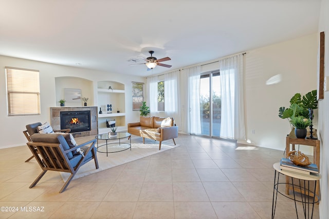 living room with ceiling fan, a tiled fireplace, light tile patterned floors, and built in shelves