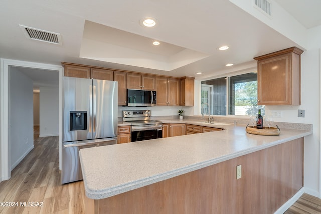 kitchen with kitchen peninsula, appliances with stainless steel finishes, sink, a tray ceiling, and light hardwood / wood-style flooring