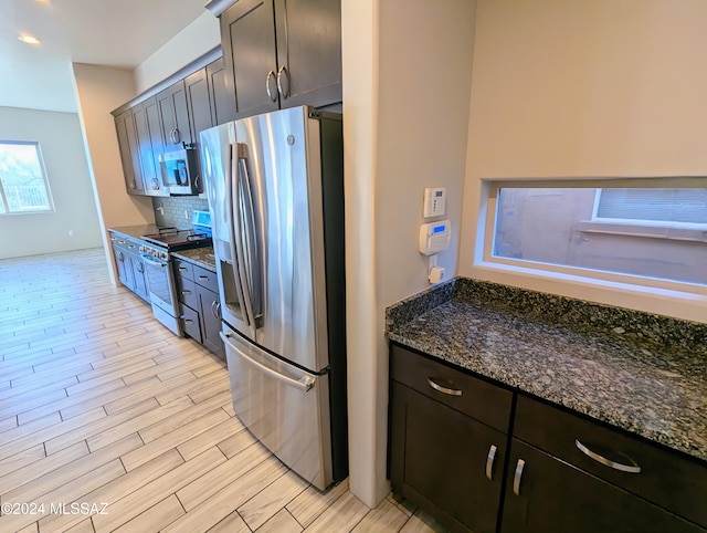 kitchen with stainless steel appliances, tasteful backsplash, dark brown cabinets, light wood-type flooring, and dark stone countertops