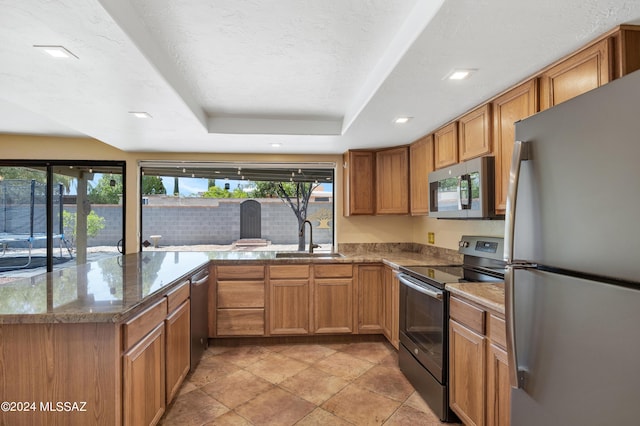 kitchen featuring sink, dark stone countertops, appliances with stainless steel finishes, a tray ceiling, and kitchen peninsula